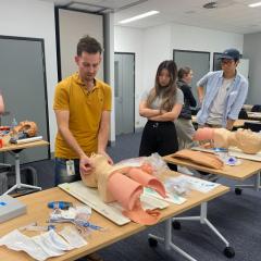 Three people standing in room watching man do CPR on Mannequin